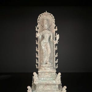 May include: A detailed, weathered statue of a standing figure with multiple arms, adorned with intricate carvings and a halo. The statue is a muted gray-green color and stands on a decorative base.