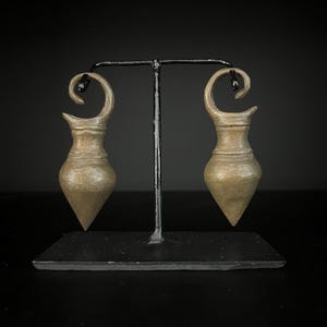 Puede incluir: Dos objetos decorativos en forma de lágrima de color bronce con una parte superior curvada, expuestos sobre un soporte de metal negro. Los objetos tienen una superficie texturizada y una parte inferior puntiaguda, sobre un fondo oscuro.