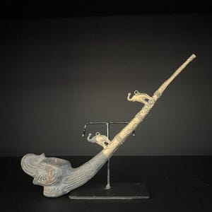 Könnte beinhalten: Eine antike Metallpfeife mit einem Elefantenkopf-Kopf und dekorativen Elefantenfiguren. Die Pfeife wird von einem schwarzen Metallständer vor einem dunklen Hintergrund getragen. Die Pfeife hat einen langen, schlanken Stiel.