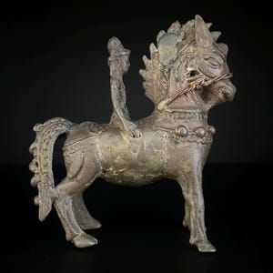 May include: A bronze-colored sculpture of a horse with a rider. The horse has detailed features, including a decorative mane and a bridle. The rider is seated upright. The sculpture is set against a dark background, highlighting its intricate design and antique appearance.