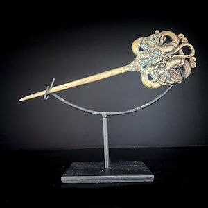 Pode incluir: Grampo de cabelo antigo com uma cabeça ornamentada de cor bronze com um design detalhado de dragão. O grampo tem um longo e fino eixo de cor marfim e é exibido em um suporte de metal sobre um fundo escuro.
