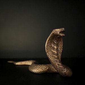 May include: A detailed, golden cobra statue with an open mouth and flared hood. The snake is posed in a striking, upright position, with intricate scale detailing. The sculpture is set against a dark background.