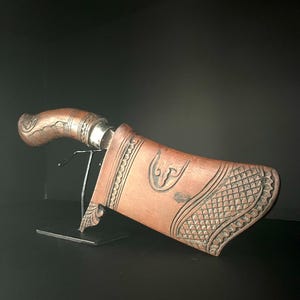 Può includere: Un coltello artigianale con manico in legno e fodero in pelle decorativo. Il fodero presenta intricati motivi intagliati e un design curvo. Il coltello è esposto su un supporto nero su sfondo scuro.