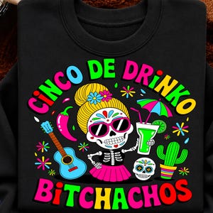Peut inclure: Un sweat-shirt noir avec un motif coloré représentant un crâne en sucre avec des cheveux blonds, des lunettes de soleil et une jupe rose. Le design inclut une guitare bleue, un cactus vert, une boisson au citron vert et le texte CINCO DE DRINKO BITCHACHOS.