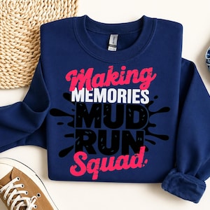 May include: A navy blue crewneck sweatshirt featuring the text "Making MEMORIES MUD RUN Squad" in a combination of pink, white, and black lettering. The word "MUD" is layered over a black ink-like splash graphic on the front of the garment.
