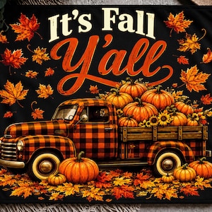 Puede incluir: Una manta rectangular negra con el texto It's Fall Y'ALL en letras blancas y naranjas. El diseño incluye un camión vintage con un patrón de cuadros naranjas y negros, rodeado de calabazas naranjas y hojas de otoño esparcidas.