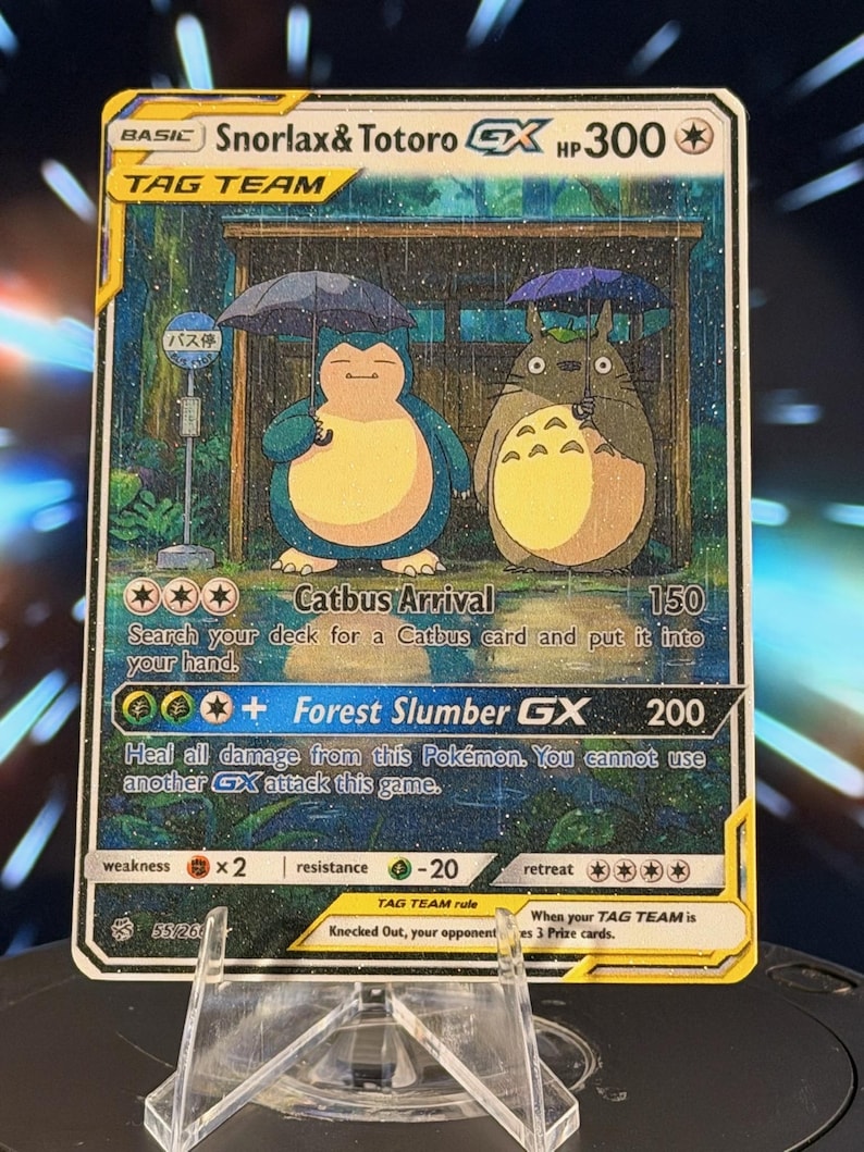 K&ouml;nnte beinhalten: Eine Pok&eacute;mon-Sammelkarte mit Snorlax und Totoro unter Regenschirmen in einer regnerischen Szene. Die Karte hat einen goldenen Rand und Text wie "TAG TEAM" und "Forest Slumber GX". Die Karte wird auf einem transparenten St&auml;nder pr&auml;sentiert.