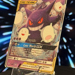 K&ouml;nnte beinhalten: Eine holografische Gengar & Warawara GX Tag Team Sammelkarte. Die Karte zeigt ein lila Gengar mit roten Augen und mehrere wei&szlig;e, geisterartige Kreaturen. Die Karte wird auf einem transparenten Acrylst&auml;nder pr&auml;sentiert.