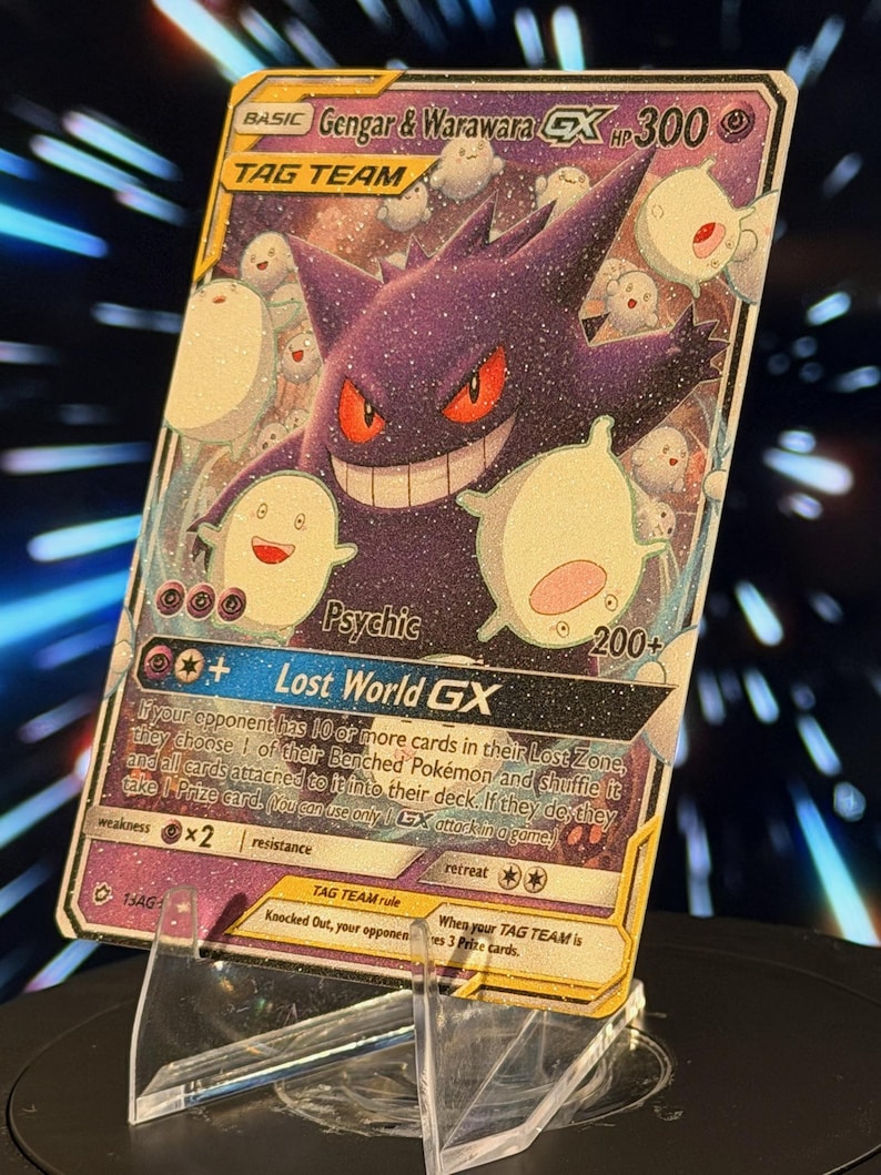 K&ouml;nnte beinhalten: Eine holografische Gengar & Warawara GX Tag Team Sammelkarte. Die Karte zeigt ein lila Gengar mit roten Augen und mehrere wei&szlig;e, geisterartige Kreaturen. Die Karte wird auf einem transparenten Acrylst&auml;nder pr&auml;sentiert.