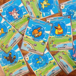 May include: A collection of colorful trading cards featuring various cartoon characters. Each card has a unique design with a blue background, character illustrations, and text. The cards are scattered, showcasing different characters and card details.
