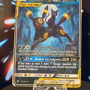 K&ouml;nnte beinhalten: Eine Pok&eacute;mon-Sammelkarte mit Umbreon und Jiji GX. Die Karte hat einen dunkelblauen Hintergrund mit den beiden Charakteren im Vordergrund. Die Karte enth&auml;lt Text, der die F&auml;higkeiten und Werte des Charakters beschreibt. Die Karte wird in einem transparenten Kunststoffhalter angezeigt.
