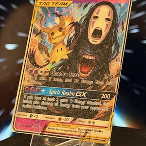 K&ouml;nnte beinhalten: Eine Sammelkarte mit Mimikyu und Ohne Gesicht. Die Karte hat einen goldenen Rand und enth&auml;lt den Text "Mimikyu & No Face GX" und "TAG TEAM". Die Karte wird auf einem transparenten Acryl-St&auml;nder pr&auml;sentiert.