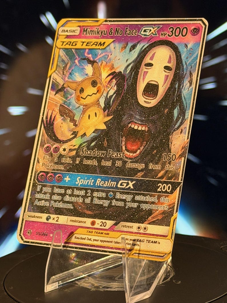 K&ouml;nnte beinhalten: Eine Sammelkarte mit Mimikyu und Ohne Gesicht. Die Karte hat einen goldenen Rand und enth&auml;lt den Text "Mimikyu & No Face GX" und "TAG TEAM". Die Karte wird auf einem transparenten Acryl-St&auml;nder pr&auml;sentiert.