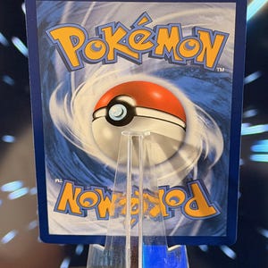 K&ouml;nnte beinhalten: Eine Pok&eacute;mon-Sammelkarte mit dem Pok&eacute;mon-Logo in Gelb und einem rot-wei&szlig;en Pok&eacute;ball-Design auf blauem Hintergrund. Die Karte wird auf einem transparenten Acrylst&auml;nder pr&auml;sentiert.
