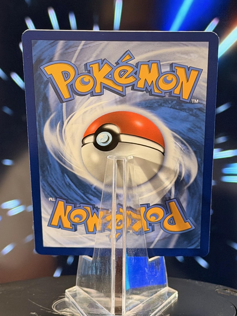 K&ouml;nnte beinhalten: Eine Pok&eacute;mon-Sammelkarte mit dem Pok&eacute;mon-Logo in Gelb und einem rot-wei&szlig;en Pok&eacute;ball-Design auf blauem Hintergrund. Die Karte wird auf einem transparenten Acrylst&auml;nder pr&auml;sentiert.
