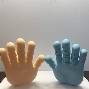 May include: Two large, stylized hand sculptures. One is a light peach color, the other is a light blue. Each hand has five fingers with rounded tips. The hands are positioned as if they are about to touch.