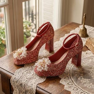 Può includere: Un paio di décolleté rosse glitterate con cinturini alla caviglia e tacchi a blocco. Ogni scarpa presenta un abbellimento floreale sulla punta. Le scarpe sono esposte su un tavolo di legno con un runner di pizzo.