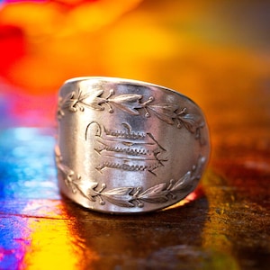 May include: A silver ring with an engraved design. The ring features a central emblem and is bordered by a laurel leaf pattern. The ring is set against a colorful, blurred background.