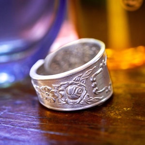 May include: A silver-toned metal ring crafted from a repurposed spoon, featuring an embossed floral pattern with a prominent rose at the center and delicate leaf details. The band has a wide, open-ended design with a textured, vintage-inspired finish.