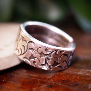 Könnte beinhalten: Ein silberner Ring mit einem eingravierten floralen Muster. Der Ring hat ein breites Band und eine leicht abgerundete Oberseite. Das filigrane Design bedeckt das gesamte Band und erzeugt ein strukturiertes Aussehen. Der Ring wird auf einer Holzoberfläche präsentiert.