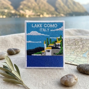May include: A rectangular embroidered patch featuring a blue sky with white clouds above a dark blue lake and a white building with yellow roofs. The text LAKE COMO ITALY is stitched in white at the top of the patch, which has a white border.
