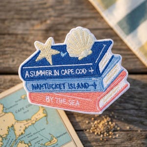 May include: An embroidered patch featuring a stack of three books with white text reading A SUMMER IN CAPE COD, NANTUCKET ISLAND, and BY THE SEA. The top book is dark blue with a yellow starfish and seashell, the middle is light blue, and the bottom is coral.