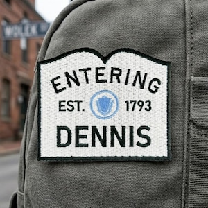 May include: A rectangular embroidered patch with a white background and black border attached to a grey canvas bag. The patch features the black text ENTERING, EST. 1793, and DENNIS, with a central light blue circular emblem containing a shield icon.