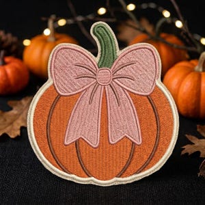 May include: An embroidered patch shaped like an orange pumpkin with a green stem and a large, light pink bow tied around the top. The pumpkin features dark orange vertical lines for texture and is outlined with a cream-colored border.