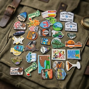 May include: A collection of various embroidered patches featuring diverse designs including national parks, landscapes, animals, and text such as Entering Cape Cod, Grand Teton, Joshua Tree, and Tahoe, arranged on a green canvas fabric surface.