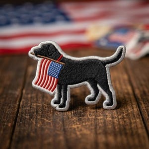 May include: An embroidered patch featuring a black Labrador retriever standing in profile. The dog holds a small American flag in its mouth, with the flag hanging down. The patch has a white stitched border and is shaped to the dog's silhouette.