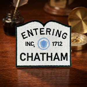 May include: A rectangular embroidered patch with a white background and black border. The text reads ENTERING, INC. 1712, and CHATHAM in black thread. A light blue circular emblem with a central shield design is positioned between INC. and 1712.