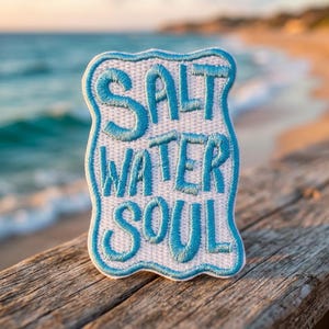 May include: A white embroidered patch with a wavy blue border and the text SALT WATER SOUL stitched in bright blue thread. The patch has a rectangular shape with rounded, scalloped edges and a textured fabric surface with visible stitching.