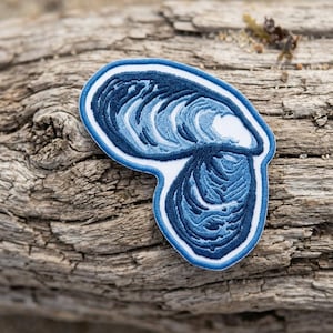 May include: An embroidered patch shaped like an oyster shell, featuring intricate thread work in various shades of blue, including navy, royal blue, and light blue, with a white border and a white interior section of the shell.