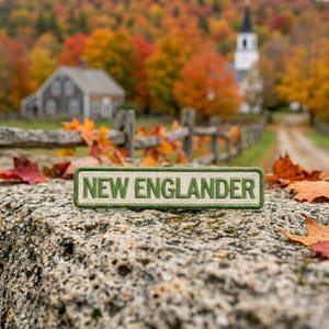 May include: A rectangular embroidered patch with a cream-colored background and a dark green border. The text NEW ENGLANDER is stitched in dark green capital letters across the center of the patch, which rests on a textured stone surface.