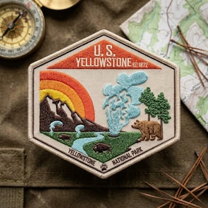 May include: A hexagonal embroidered patch featuring a landscape of Yellowstone National Park. The design includes a sunset, mountains, a geyser, two bison, a brown bear, and pine trees. Text reads U.S. YELLOWSTONE EST 1872 and YELLOWSTONE NATIONAL PARK.