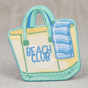 May include: An embroidered patch shaped like a tote bag in cream and yellow with mint green handles and trim. The bag features the text BEACH CLUB in blue embroidery and a blue and white striped towel draped over the side of the bag.