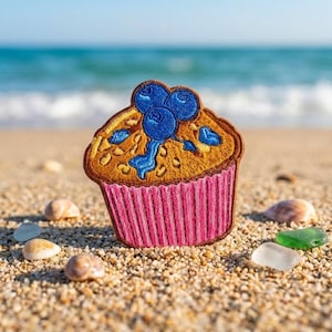 May include: An embroidered patch shaped like a muffin with a golden-brown top, blue berries, and a pink pleated paper liner. The design features detailed stitching in shades of brown, blue, and pink, creating a textured appearance for the baked good.