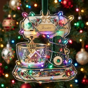 May include: A decorative Christmas ornament shaped like a stand mixer with a clear bowl containing a miniature gingerbread house, pine trees, and a deer. The mixer features the name Jessica, a base with a train, and colorful string lights.