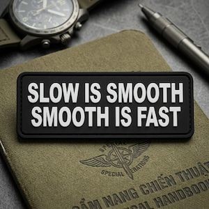 May include: A black rectangular patch with the white text "SLOW IS SMOOTH SMOOTH IS FAST." The patch is on a green notebook with a watch and pen in the background. The notebook has text and a logo.
