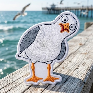 May include: An embroidered patch featuring a cartoon seagull with white feathers, dark gray wings, and bright orange webbed feet. The seagull has a surprised expression with wide eyes and an open orange beak, outlined in white thread.