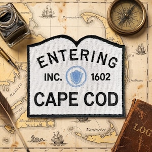 May include: A white embroidered patch with a black border featuring the text ENTERING INC. 1602 CAPE COD in black lettering. A small light blue circular emblem with a shield icon is centered between the text. The patch has a curved top edge.