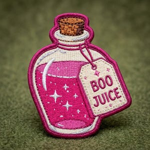 May include: An embroidered patch shaped like a potion bottle with a cork stopper. The bottle contains bright pink liquid with white star accents. A cream-colored tag is attached to the neck, featuring the text BOO JUICE in pink embroidery.