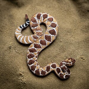May include: An embroidered patch shaped like a coiled rattlesnake. The snake features a pattern of dark brown and tan diamond-shaped scales with white outlines. The tail end shows a distinct black and white segmented rattle, while the head is small.