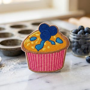 May include: An embroidered patch shaped like a muffin with a golden-yellow top, dark blue blueberries, and a pink and white striped paper liner. The design features detailed stitching in shades of yellow, blue, brown, and pink on a fabric base.