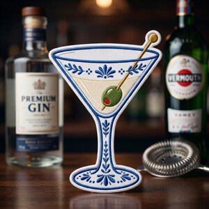 May include: An embroidered patch shaped like a martini glass with blue and white floral patterns, containing a pale yellow liquid and a green olive on a gold toothpick. The patch is positioned between a bottle of premium gin and a bottle of vermouth.