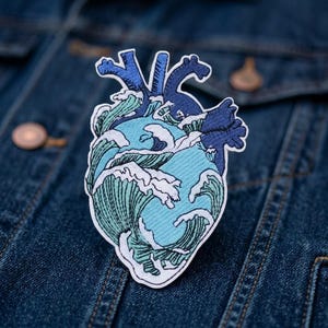 May include: An embroidered patch shaped like an anatomical heart, featuring a design of crashing ocean waves in shades of light blue, teal, and white, with dark blue vessels at the top, all outlined in a white border against a denim fabric.