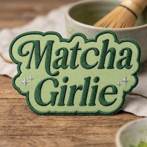 May include: A light green embroidered patch with a dark green scalloped border. The text "Matcha Girlie" is stitched in dark green cursive lettering. Two small, silver-colored four-pointed star icons are positioned on the left and right sides.