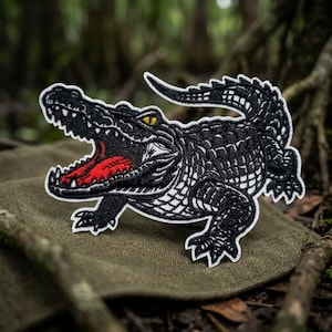 May include: An embroidered patch featuring a black alligator with a wide-open mouth, sharp white teeth, a bright red tongue, and a single yellow eye. The design is outlined in white thread, showcasing detailed scales and a curved tail.