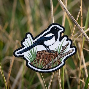 May include: An embroidered patch featuring a black and white magpie perched on a brown tree stump. The bird has a long tail, white belly, and black wings. Green grass blades and a small wooden branch are depicted around the base of the stump.