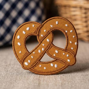 May include: A brown pretzel-shaped embroidered patch featuring a textured golden-brown thread base and small, scattered white embroidered dots representing salt crystals. The patch has a distinct stitched border and a classic knotted loop design.